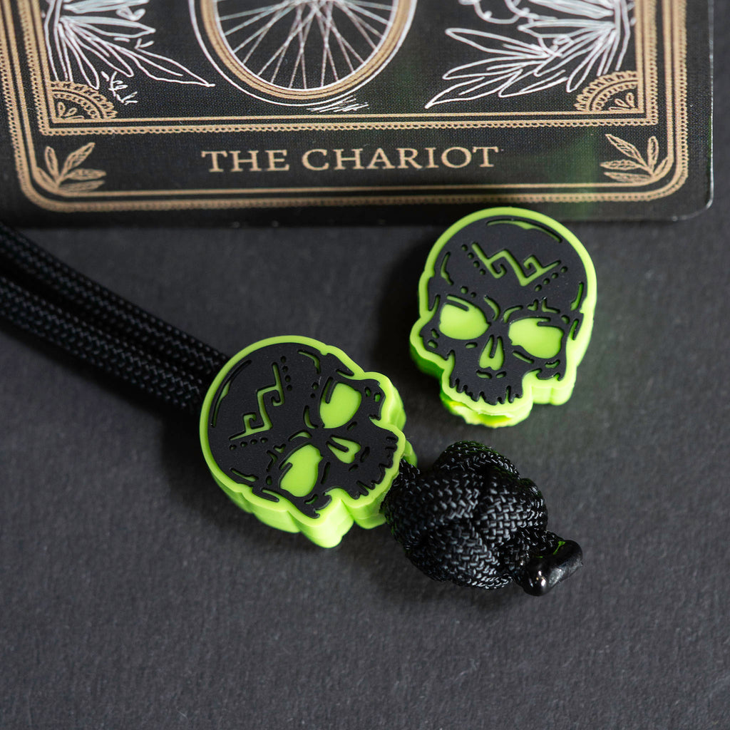 Two neon green skull-shaped beads with black cutout details, attached to a black braided cord, displayed on a dark surface with a tarot card labeled The Chariot in the background.