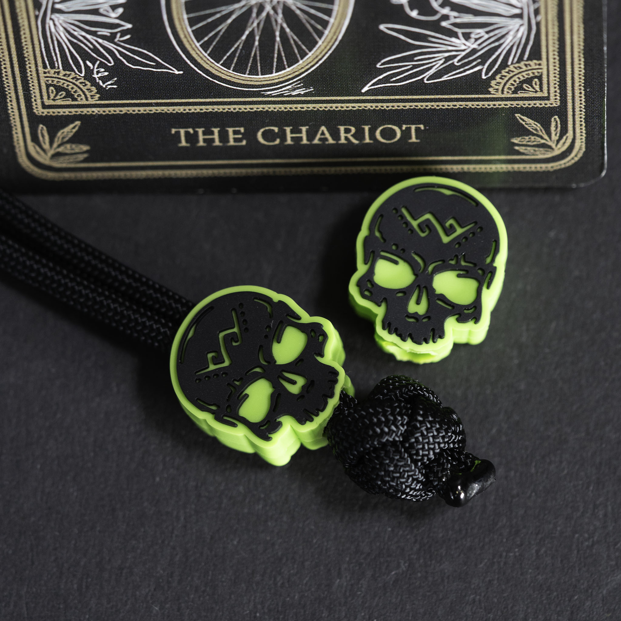Two neon green skull-shaped beads with black cutout details, attached to a black braided cord, displayed on a dark surface with a tarot card labeled The Chariot in the background.