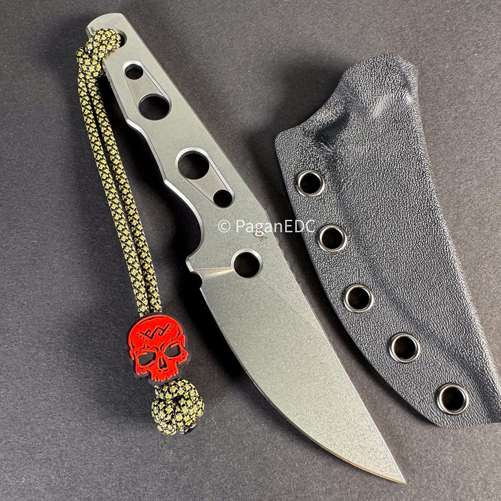 Compact fixed-blade EDC knife with a skeletonized gray stonewashed steel handle and matching stonewashed blade, shown beside a black Kydex sheath with eyelets. The knife features multiple handle cutouts, a straight cutting edge with pointed tip, and a braided paracord lanyard finished with a red skull bead, all photographed on a dark background.