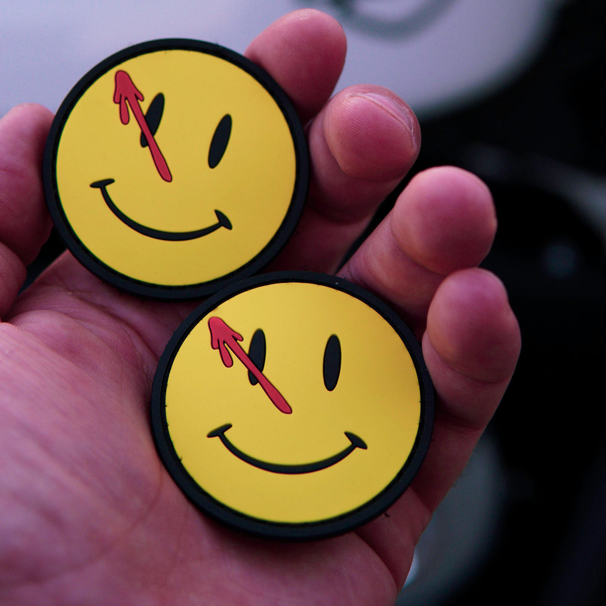 Two Watchmen PVC morale patches with yellow smiley faces and red blood splatter held in a hand